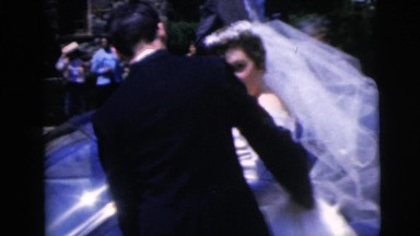 1962: groom prepares to open the car door for his bride as the wind catches her