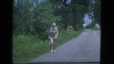 1967: an older woman wearing a light brown dress walking down the road. NEVADA