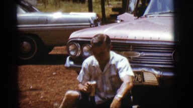 1968: man sitting on ground in front of car takes relaxed sip of canned beverage