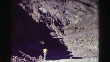 1969: woman in yellow shirt and blue jeans sits on rock near mountain path