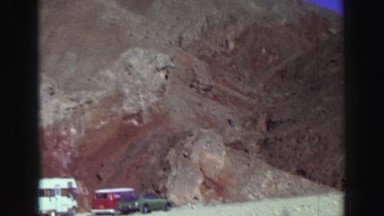 1969: cars parked under sunny mountain, person walks toward shadowy cleft