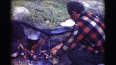 1951: man prepares food near camp fire before standing and tossing