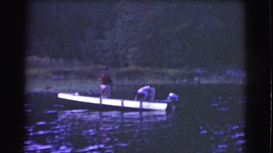 1951: two men on a small metal motor boat in the water just off the shoreline