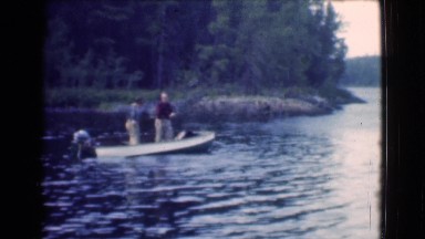 1951: two men on a small metal motor boat in the water just off the shoreline