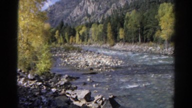 1967: train runs along the edge of river near wooded area. COLORADO