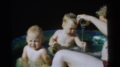 1961: man sits in a small shallow pool with two babies and pours water out into