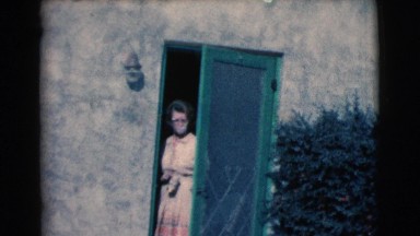 1961: an older lady in a tan dress and glasses goes to, and opens the door