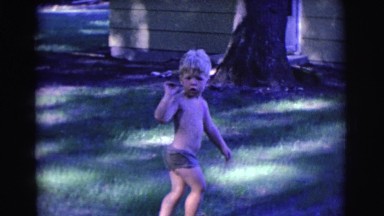 1961: a shirtless young boy in shorts looks confused as he walks in his yard