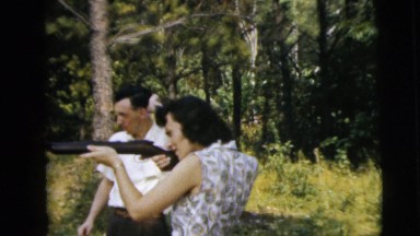 1962: a woman with aiming a long gun in the woods as a man looks on CAMDEN