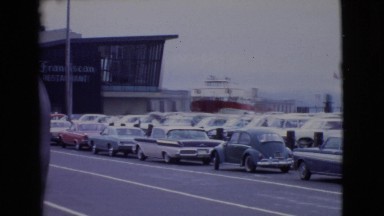 1968: old woman walking on the pedestrian path near to the car parked area