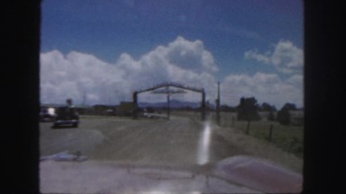 1958: a car is being driven on a dirt road through a rural open gateway.