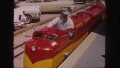 1958: red and yellow toy train pulls away from platform as lady walks
