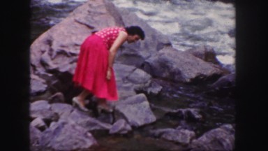 1958: hiking women in formal red dress sits by fast flowing river water COLORADO