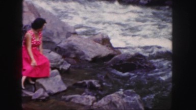 1958: women lounging in formal red dress sits by fast flowing river water