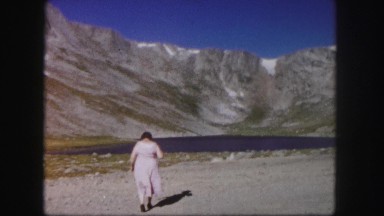 1958: stout woman in pink dress strides away towards lake with hills