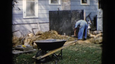 1960: man in jeans and grey shirt pottering about a junk-strewn yard measuring