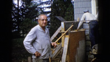 1960: man smoking cigarette watching two workers rebuilding outside stairway.