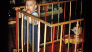 1959: a baby in overall peering over the railing of a crib like enclosure