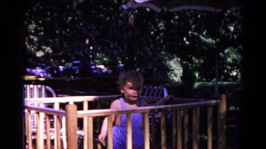 1963: toddler wearing blue romper standing inside playpen placed in a yard.