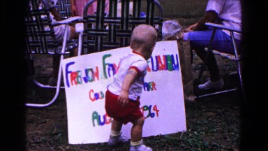 1963: a toddler stands in front of a handwritten sign, seated adults and a girl.