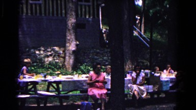 1963: adults and children are gathered at picnic tables by a rustic building.