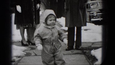1948: a child dressed in a skiing suit is walking on the sidewalk amongst other