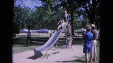 1964: a little boy takes a turn on a slide in the park while adults watch.