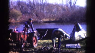 1962: two men working outdoors with a tractor near boats on the water's edge