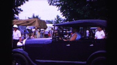 1962: people gathered in a park with tents, with a woman driving antique car
