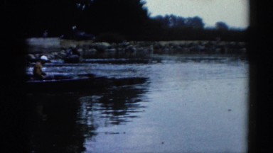 1962: two men on a small metal motor boat in the water just off the shoreline