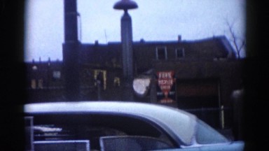 1962: driving past a town block passing cars, pedestrian and buildings.