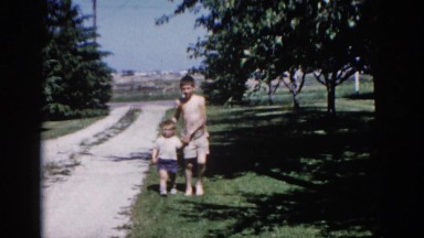 1962: older boy guides younger boy along the edge of a lawn by a driveway.