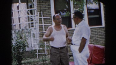 1962: two middle-age men stand in front of house and have a conversation in yard
