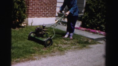 1956: a lady in blue shirt and blue jeans trying to start a lawn mower in yard.