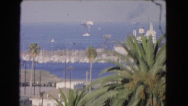 1966: sunny day on a beach with boats docked on the ocean as a small plane flies
