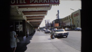 1966: lady walks down a sidewalk in a city on a busy street while cars pass by