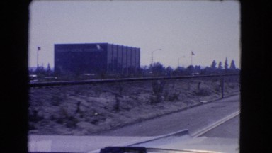 1966: a car is moving on top of a highway lane passing traffic and a building.