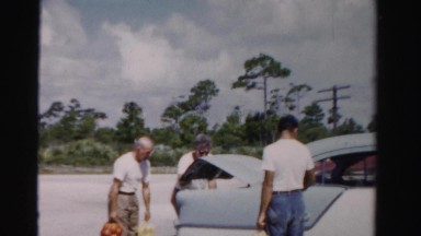 1955: three men pack the trunk of an antique, light blue car by the highway.