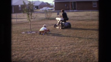 1975: Father Pulling Child in Wagon Behind Lawn Mower California