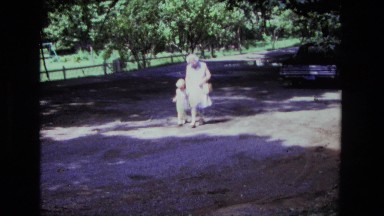 1975: woman in her seventies strolling with young child down an unpaved road in