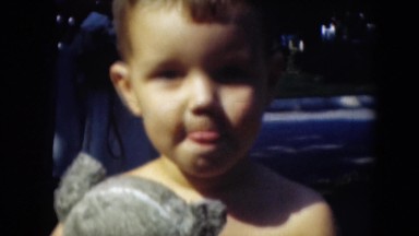 1947: boy holding teddy bear stares into camera while countryside golfer tees