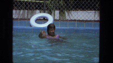 1976: girl in pink bathing suit splashing and playing in fenced swimming pool