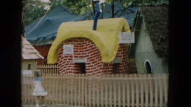 1957: girl peeking at the miniature house park. pigs feeding in dirty bowl.