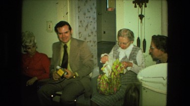 1974: older woman opening up wrapped gift as she is watched by family