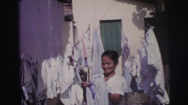 1967: smiling lady hanging clothes out to dry near house in the warm summer