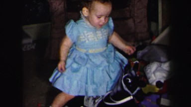 1972: little girl in blue dress sitting on toy horse and pile of other toys