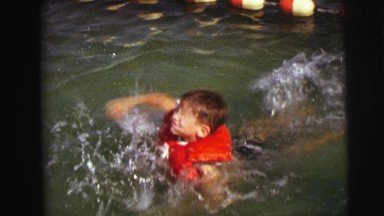 1974: young boy learning to swim by paddling in pool with adults