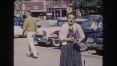 1962: woman in dress and glasses talking waving near parked antique cars