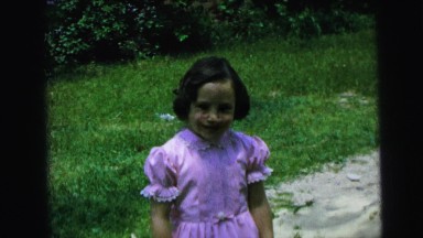 1964: a small girl smiles in a fancy pink dress on a nice day in the yard.