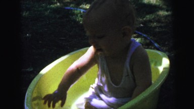 1964: a toddler outdoors playing in a yellow bucket full of water CAMDEN
